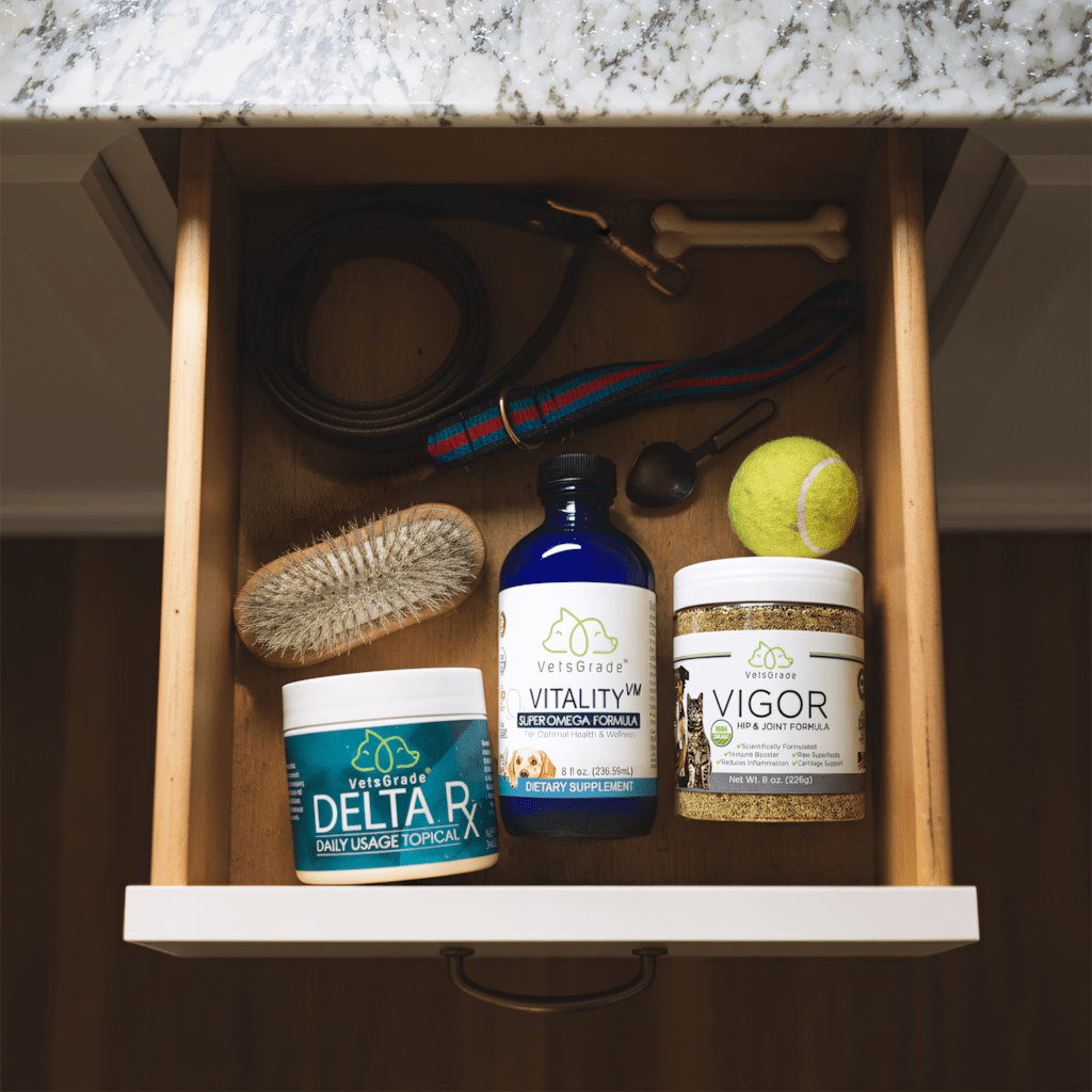 VetsGrade Senior Pack products in a kitchen drawer — Delta RX topical balm, Vitality VM omega-3 bottle, and Vigor hip and joint powder alongside a dog leash, brush, and tennis ball